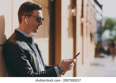 Young side view fashionable man in black leather jacket shirt glasses stand near building wall outdoors, talk by mobile cell phone use air pods headphones Concept of people urban lifestyle and clothes - Powered by Shutterstock