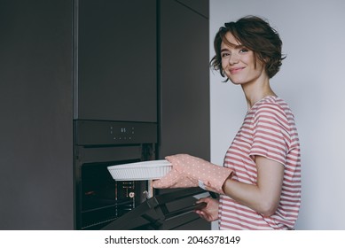 Young Side View Housewife Woman In Casual Clothes Striped T-shirt Puts Casserole In Baking Dish In Oven Cooking Look Camera Food In Light Kitchen At Home Alone. Healthy Diet Bakery Lifestyle Concept.