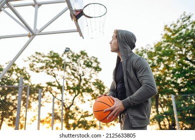 Young Side View Sporty Sportsman Man 20s Wearing Grey Sports Clothes Hood Training Holding In Hand Ball Shooting Free Throw Play At Basketball Game Playground Court. Outdoor Courtyard Sport Concept.