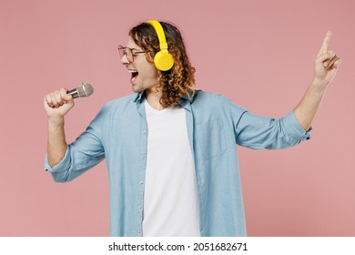 Young Singer Expressive Man 20s With Long Curly Hair Wearing Blue Shirt White T-shirt Glasses Headphones Sing Song In Microphone Isolated On Pastel Plain Pink Color Wall Background Studio Portrait.