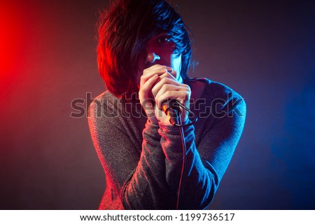 The young singer or vocalist in emo style is singing on concert on background of red and blue concert lights.