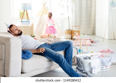 Young Single Father Overwhelmed With Household Chores Sleeping On Sofa In Messy Living Room With His Little Daughter Playing In The Background. 