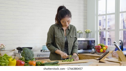 Young Single Lady In Modern Home Kitchen Cooking Food For Dinner. Asian Chinese Woman Cutting Vegetables By Knife On Wooden Board. Girl Standing In City Apartment Chopping Cucumber For Healthy Meal.