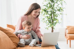 Young Single Mother Mom With Toddler Newborn Baby Infant Son Using Laptop, Searching Web, Watching Cartoons Together. Working From Home On Maternity Leave, Freelancer, Looking For Kids Goods