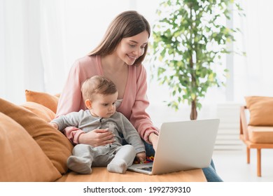 Young Single Mother Mom With Toddler Newborn Baby Infant Son Using Laptop, Searching Web, Watching Cartoons Together. Working From Home On Maternity Leave, Freelancer, Looking For Kids Goods