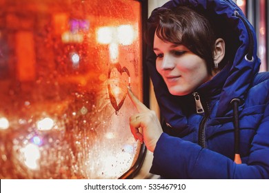 Young single woman in blue jacket sitting in an empty tram and paints on glass heart