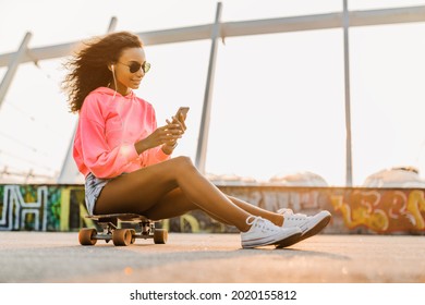 Young skateboard girl using phone and earphones outdoors. African woman communicating with friends, typing messages, texting online on cellphone close to stadium. - Powered by Shutterstock