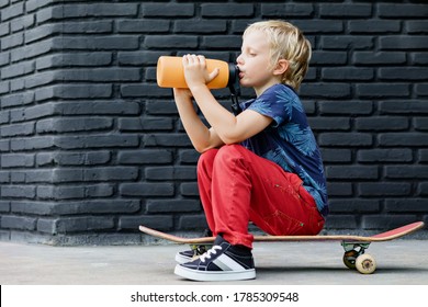 Young Skater Sit On Skateboard, Drink Fresh Water From Reusable Bottle After Children Training Class. Active Family Lifestyle, Outdoor Recreational Activities On Summer Holidays In City. Kids Sports.