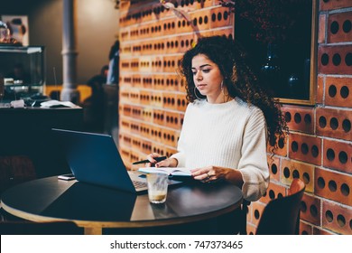 Young Skilled Freelancer Watching Training Webinar In Webpage While Sitting In Co-working Space.Pondering Cute Female Student Preparing For Upcoming Examination Using Laptop Computer And Notepad