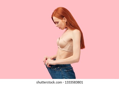 Young Skinny Anorexic Woman With Anxious, Worried In Oversized Jeans Showing Her Diet Results On Pink Background