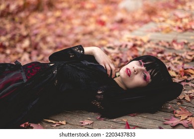 Young And Skinny Hispanic Goth Girl With Black Dress, Cross, Sexy Face And Red Makeup Lying In A Wood Platform In The Autumn Forest With Red, Orange And Yellow Dry Leaves