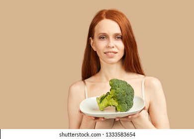 Young skinny woman with ginger hair and starved face in underwear looking at camera holding plate with raw broccoli, happy to eat something