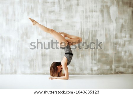 Young skinny woman in grey leotard doing yoga handstand in light grey studio