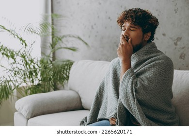 Young sleepy exhausted tired sick ill Indian man wapped in plaid yawn cover mouth with hand sits on grey sofa couch stay at home hotel flat spend free spare time in living room indoor Lounge concept