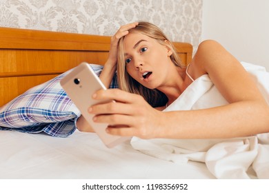 A Young Sleepy Girl In Shock, With Her Eyes Wide Open In Her Mouth, Lying In Bed, Looks At The Smartphone And Realizes That She Is Late For The Robot