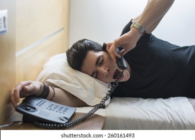 Young Sleepy Man, Lying On His White Bed, Yawning While Talking To Someone Through The Telephone