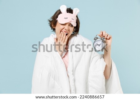 Young sleepy woman in pajamas jam sleep eye mask rest relaxing at home wrap cover under blanket duvet hold clock alarm yawning isolated on pastel blue background studio Bad mood night bedtime concept