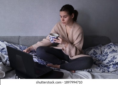 Young Sleepy Woman In Sweatpants And A Pink Cardigan Sits In Bed In Front Of A Laptop With A Cup Of Coffee. Relaxed Remote Work. Communication Via The Internet. Video Communication