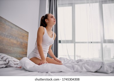 若いほっそりした白人女性が音楽を聴き、ヘッドフォンでベッドに座っている。 パジャマで