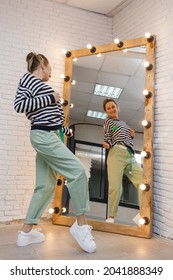 A young slender fashionable сaucasian woman in a green pants and striped jumper tries on an outfit looking in a mirror with light bulbs in a clothing store. Shopping and fashion concept. Vertical shot