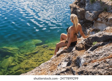 A young slender girl in a black bikini sunbathes on the rocky shore of a lake on a summer day.  - Powered by Shutterstock