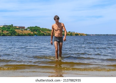 A young slender man in swimming trunks and sunglasses walks out of the river on Strilka beach in Mykolaiv, Ukraine. Rest of people at the confluence of the rivers Inhul and Pivdennyi Buh