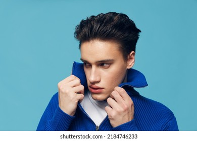 a young and slender man with thick black hair standing on a blue background in a blue zip-up sweater with a white T-shirt under it.Horizontal studio shot.
