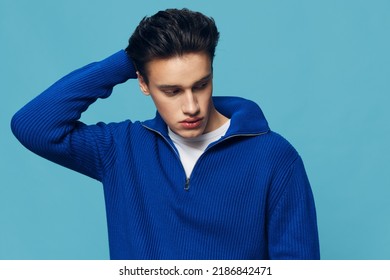 a young and slender man with thick black hair standing on a blue background in a blue zip-up sweater with a white T-shirt under it.Horizontal studio shot.