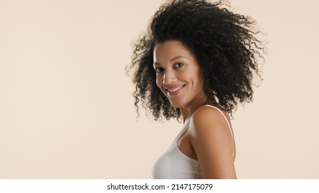 Young Slim African American Woman With Curly Hair In White Top Looks At Camera And Smiles On Beige Background | Hair Care Products Commercial