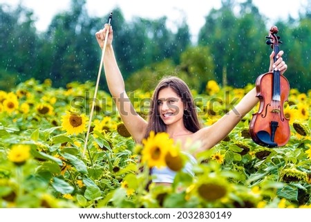young slim brunette with long hair standing in sunflower field and playing violin in rainy day