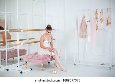 Young Slim Dancer Puts On Pointe Shoes, Sitting On A Ottoman - Bench. Photo In The Dance Hall.