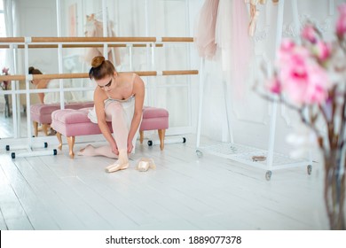 Young Slim Dancer Puts On Pointe Shoes, Sitting On A Ottoman - Bench. Photo In The Dance Hall.