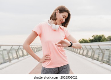 Young Slim Female Athlete Sportswoman Checking Time On Her Wrist Watch In Fitness Clothes Preparing For Jogging Running On A City Bridge In Urban Area. Active Lifestyle Concept