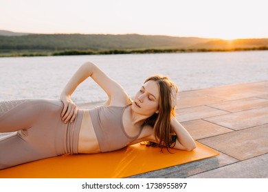 Young Slim Fit Woman On A Sport Mat In Yoga Side Lying Pose With Leg Lift, At The Sunset, Outdoors. Pilates Teacher Doing Exercises.