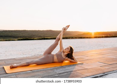 Young Slim Fit Woman On A Sport Mat In Yoga Side Lying Pose With Leg Lift, At The Sunset, Outdoors. Pilates Instructor Doing Exercises.