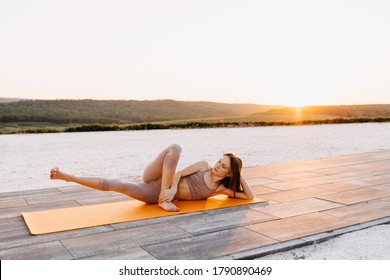 Young Slim Fit Woman On A Sport Mat In Yoga Side Lying Pose With Leg Lift, At The Sunset, Outdoors. Pilates Instructor Doing Exercises.