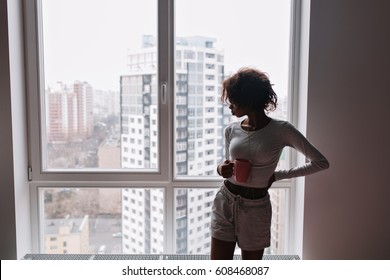 Young slim girl, teen with cup of coffee, tea looking in big window in room, high-rise buildings on background, morning in city. She has short curly hair. Wearing  long sleeves t-shirt, shorts. 