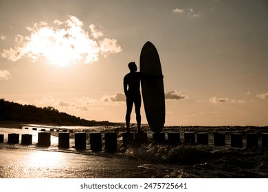 Young slim male surfer in wetsuit looking at his surfboard while standing on wooden stumps against sunset background - Powered by Shutterstock