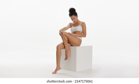 Young slim pretty African American woman with curly hair in bun in white underwear strokes smooth leg sitting on white cube platform on white background | Skin care concept - Shutterstock ID 2147175061