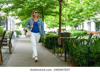 Young slim smiling woman in white pants and blue blouse and denim jacket walking down the street among gardens of cafes and restaurants in city center on sunny day. Everyday life in city