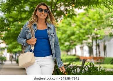 Young slim smiling woman in white pants and blue blouse and denim jacket walking down the street among gardens of cafes and restaurants in city center on sunny day. Everyday life in city