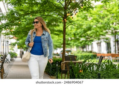 Young slim smiling woman in white pants and blue blouse and denim jacket walking down the street among gardens of cafes and restaurants in city center on sunny day. Everyday life in city