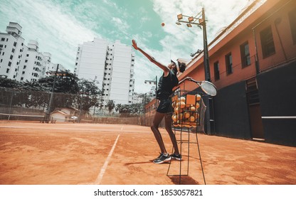 A young slim sport African-American woman is having a warm-up session on a tennis court, she's hitting tennis balls one by one taking them from the basket in the foreground, a warm sunny day - Powered by Shutterstock