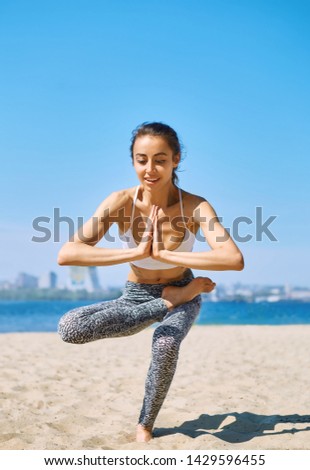 Young slim sports woman makes yoga balance exercises on the sand beach with city background against blue sky and sea. Young pretty fitness lady mixed race of Asian Caucasian ethnicity outdoors on the