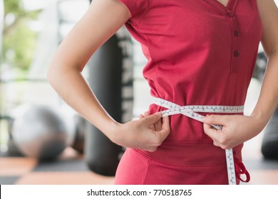 Young Slim Woman Measuring Her Waist By Measure Tape After A Diet With Accessory In Sporty Gym As Background.