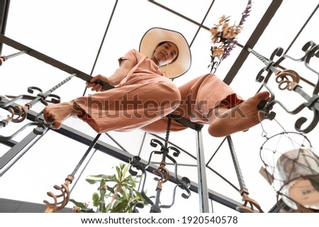 young slim woman sitting on a railing inside a building with bare feet, wears casual clothes with straw hat, perspective from below