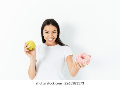 Young slim woman smiles and holds out a sweet donut instead of a green apple, the concept of a balanced diverse diet, white background - Powered by Shutterstock