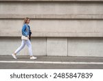 Young slim woman in white pants and denim katana walking with black bag along the sidewalk in the city against the background of gray building. Everyday life in city. Side view.