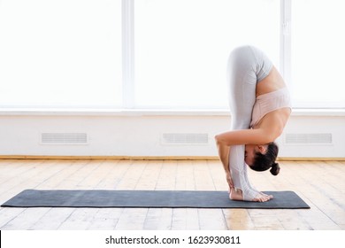 Young slim woman yoga instructor does head to knees exercise uttanasana standing forward bend pose in spacious room with large panoramic window. Concept of yoga and stretching. Advertising space
