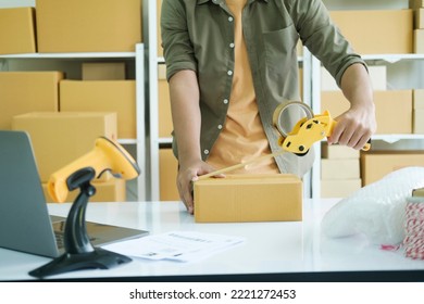 Young Small Business Entrepreneur Man, Online Store Owner With Shelves Of Boxes In The Background Packing Product In Mailing Box Sealing Box With Tape At Workplace For Shipping To Customer.